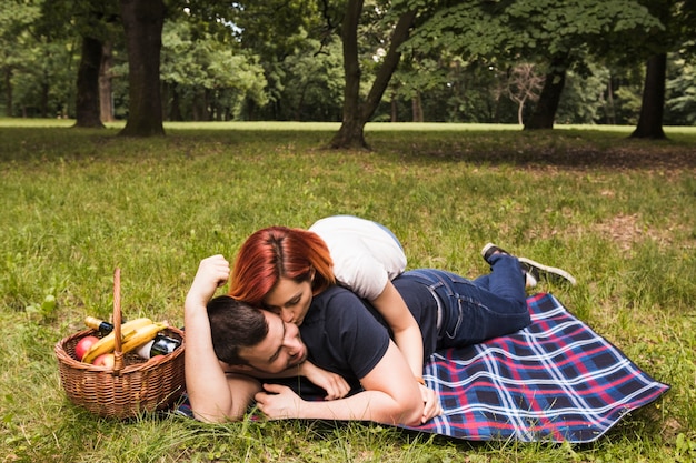 Mujer que besa a su novio tendido en una manta sobre la hierba verde en el parque