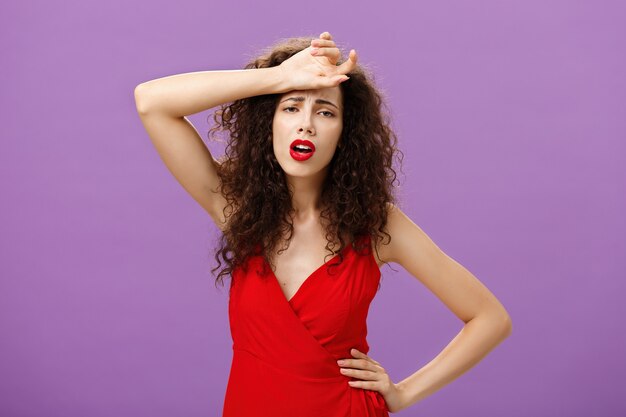 Mujer que se desmaya sintiéndose mal azotando el sudor de la frente de pie agotada y drenada sobre fondo púrpura en un elegante vestido rojo que expresa sentimientos sombríos e infelices que desean ayuda. Copia espacio