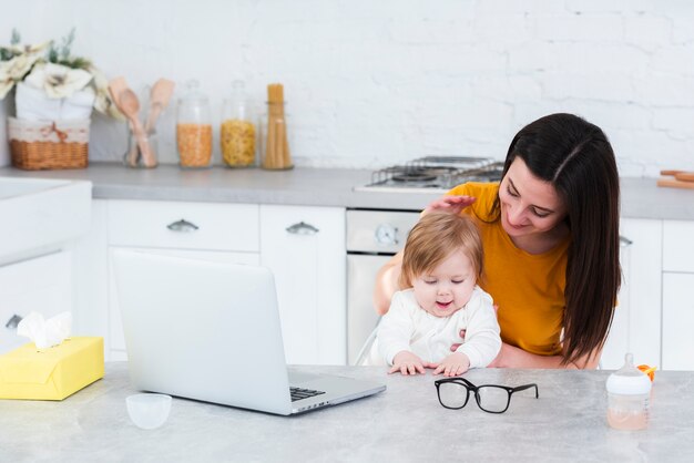 Mujer que detiene al bebé en la cocina con la computadora portátil