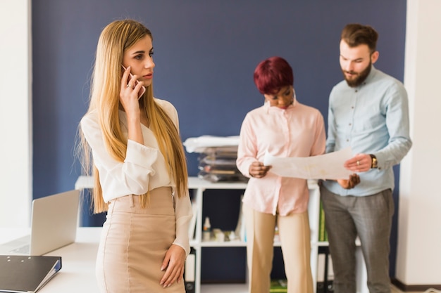 Mujer que habla en el teléfono inteligente cerca de colegas que trabajan