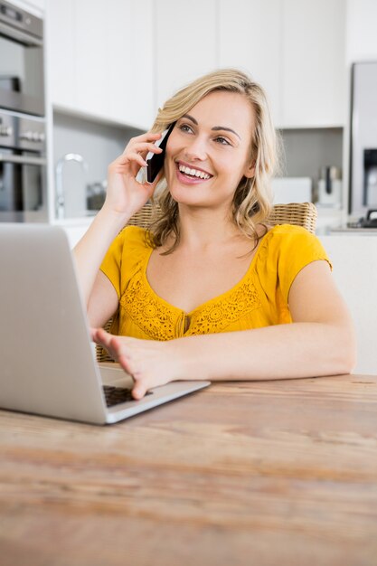 Mujer que habla en el teléfono móvil con el ordenador portátil en la cocina
