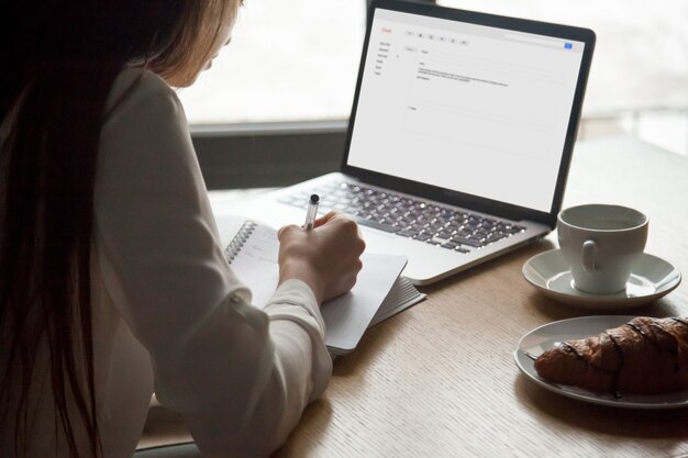 Mujer que hace notas que lee la letra del correo electrónico en el ordenador portátil en café