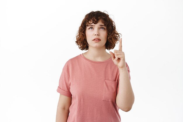 mujer que levanta las cejas vacilante, apuntando y mirando hacia arriba con la cara cuestionada dudosa, leyendo el espacio de la copia en la parte superior, de pie con una camiseta en blanco.