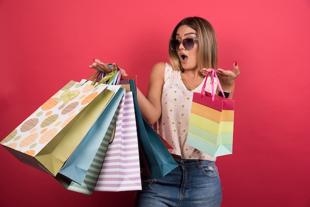 Foto gratuita mujer que lleva bolsas de la compra con expresión de sorpresa en la pared roja.