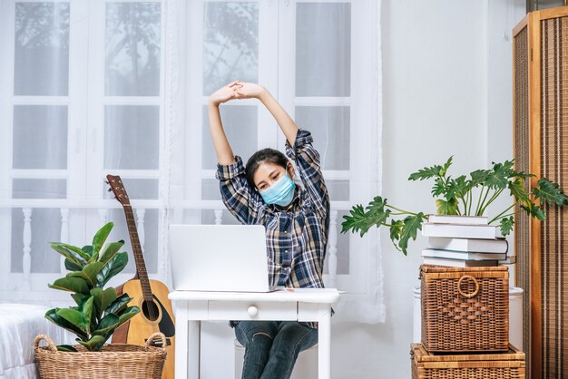 Una mujer que lleva una máscara usa una computadora portátil en el trabajo y estira los brazos para relajarse.