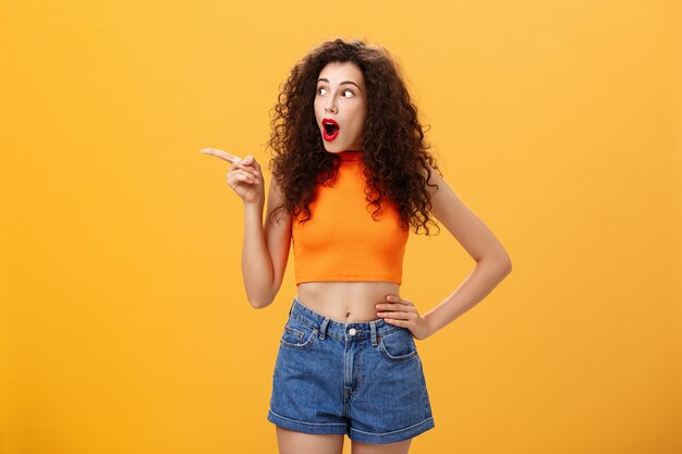 Mujer que mira a escondidas en la ventana de los vecinos que está sorprendida y sorprendida señalando y mirando fijamente sin palabras dejó de pie sobre un fondo naranja con la mandíbula caída mirada intrigada y emocionada en la parte superior recortada.