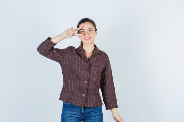mujer que muestra el signo V cerca del ojo en camisa, jeans y luciendo alegre. vista frontal.