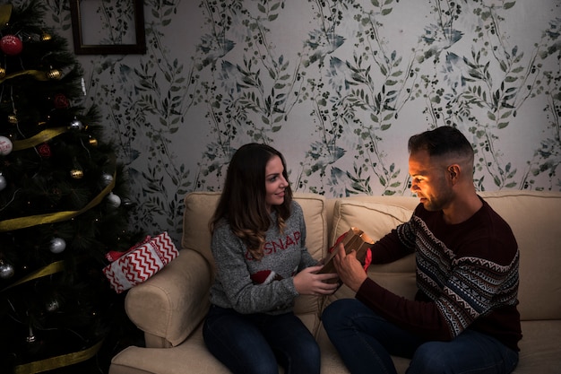 Mujer que presenta el paquete de regalo para el hombre en el sofá cerca del árbol de Navidad