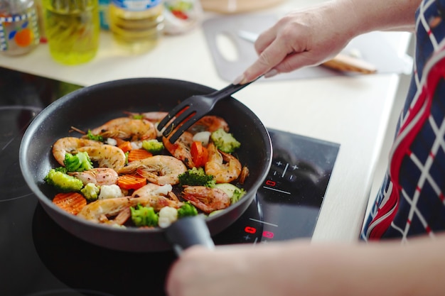 Mujer que se queda en la cocina de casa y cocinar camarones con verduras en la sartén. Concepto de cocina casera o cocina saludable