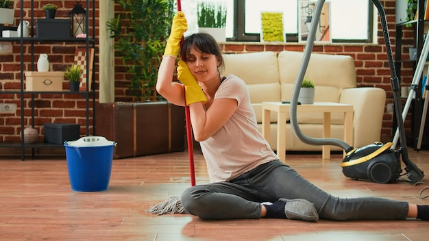 Mujer que se siente cansada después de terminar la limpieza de primavera, sentada en un suelo de madera limpio con fregona y guantes. Joven mujer soñolienta orgullosa de un apartamento ordenado, las tareas domésticas terminaron.