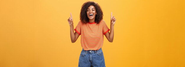 Mujer que se siente divertida y entretenida Retrato de una feliz y elegante chica afroamericana con peinado afro riéndose a carcajadas alegremente apuntando hacia arriba con los brazos levantados sobre la pared naranja