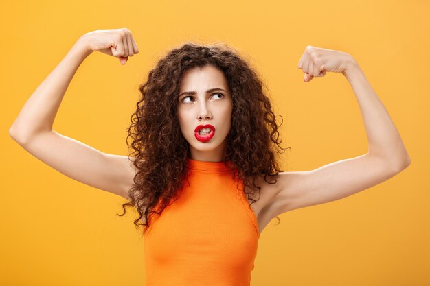 Mujer que se siente poderosa y fuerte levantando las manos con los puños cerrados haciendo que la cara intensa se esté ejercitando en el gimnasio mostrando los músculos y bíceps mirando la esquina superior derecha posando sobre fondo naranja.