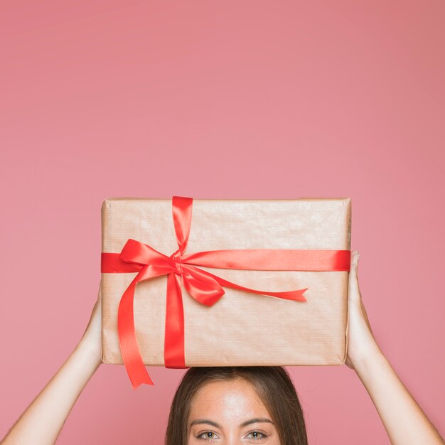 Mujer que sostiene la caja de regalo envuelta sobre su cabeza contra fondo rosado