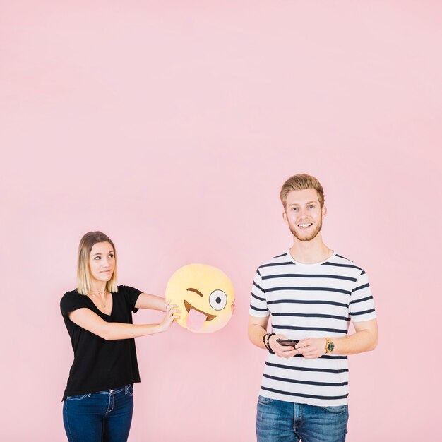 Mujer que sostiene el icono de emoji guiño cerca de hombre sonriente con teléfono móvil