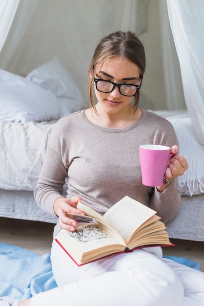 Foto gratuita mujer que sostiene el libro de lectura rosado de la taza de café en el dormitorio