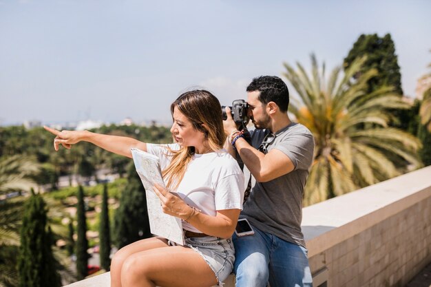 Mujer que sostiene el mapa que señala el dedo que se sienta con su novio que toma imagen en cámara en el parque