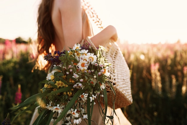 Mujer que sostiene el ramo de los wildflowers en el bolso de la paja, caminando en campo de flor en puesta del sol.