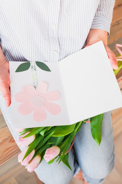 Mujer que sostiene la tarjeta de felicitación y flores rosadas