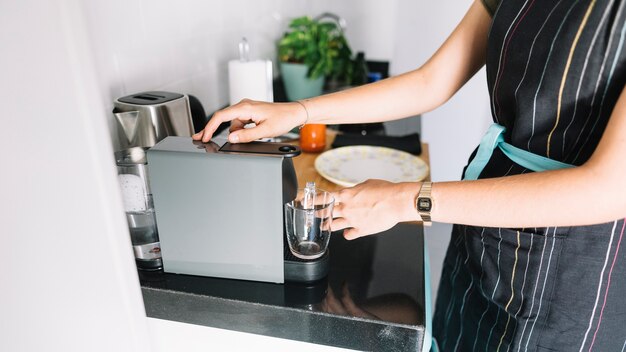 Mujer que sostiene la taza de cristal debajo de la máquina de café en cocina