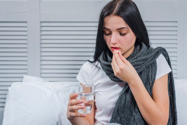 Mujer que sufre de frío sosteniendo vaso de agua tomando medicina