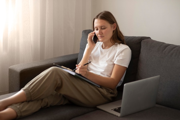 Mujer que trabaja en casa en el sofá durante la cuarentena con smartphone y portátil