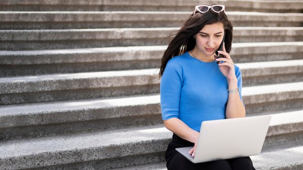 Mujer que trabaja en la computadora portátil al aire libre en pasos con smartphone