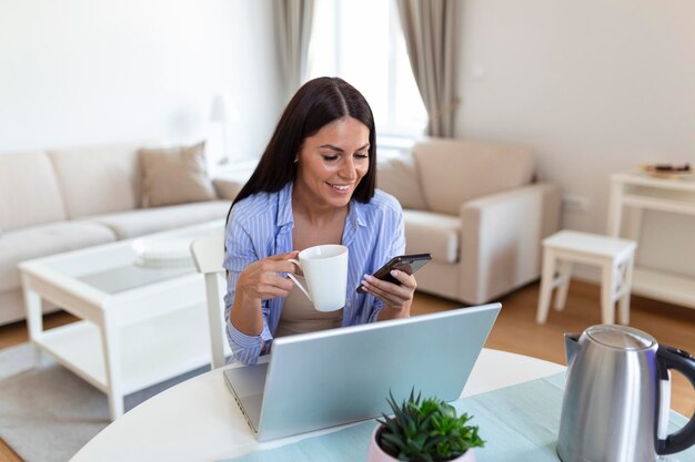 Mujer que trabaja por cuenta propia trabajando con su teléfono y computadora portátil en casa con una taza de café Mujer feliz usando un teléfono móvil mientras trabaja en casa con una computadora portátil Mujer sonriente enviando mensajes con un teléfono inteligente