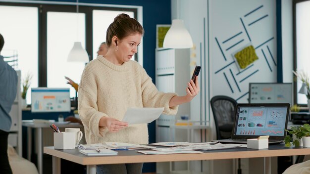 Mujer que usa un teléfono inteligente en una videoconferencia hablando de ventas con papel con gráficos en una oficina de inicio ocupada. Empleado de negocios hablando con un compañero de trabajo remoto usando un teléfono móvil.