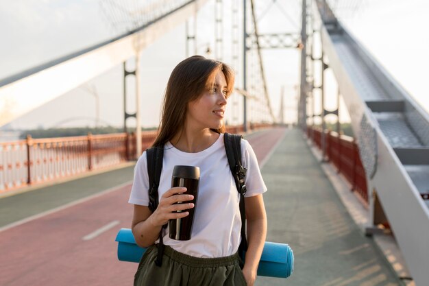 Mujer que viaja con mochila posando en el puente mientras sostiene el termo