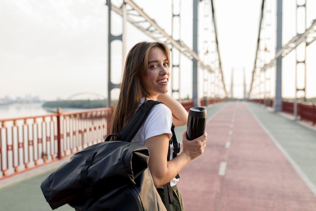 Mujer que viaja sonriente con mochila sosteniendo termo en puente