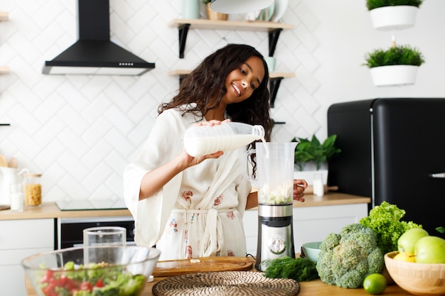 Una mujer de raza mixta bonita sonrió vertiendo leche en la licuadora cerca de la mesa con verduras frescas en la moderna cocina blanca vestida con ropa de dormir con cabello suelto