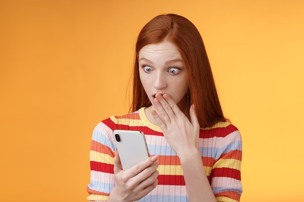 Mujer recibe impactante mensaje jadeando tapa boca palma mirando la pantalla del teléfono inteligente descubrió quién sigue a su novio en la red social de Internet de pie asombrado emocionado, fondo naranja.