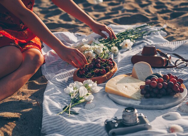 Mujer recogiendo cerezas en placa de madera con cámara vintage, flores, queso y frutas en la playa