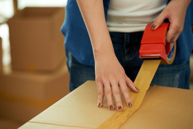 Mujer recortada irreconocible que empaca una caja con dispensador de cinta adhesiva