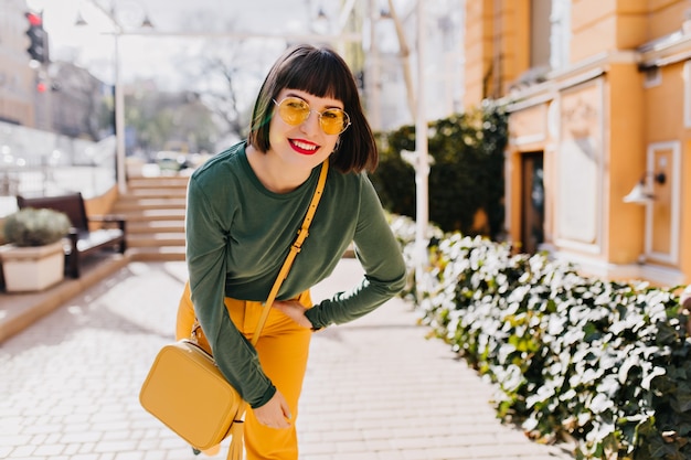 Mujer refinada con maquillaje brillante posando en la calle con gafas de sol amarillas. Tiro al aire libre de encantadora chica caucásica con cabello negro riendo en buena mañana de primavera.