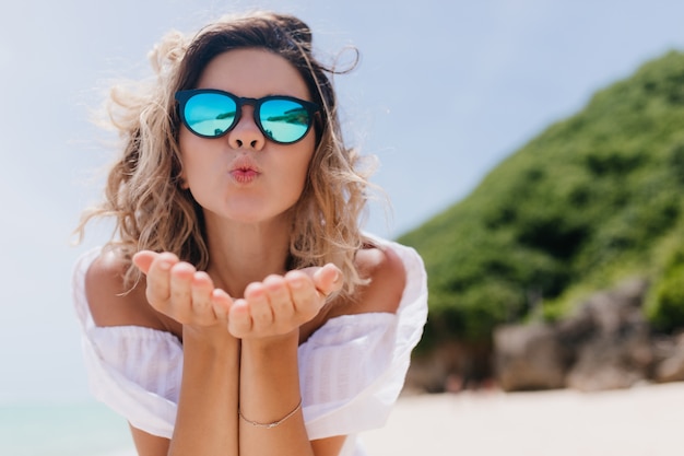 Mujer refinada con piel bronceada posando con expresión de cara de besos en la naturaleza. Disparo al aire libre del encantador modo femenino con pie de cabello ondulado en la playa en una mañana soleada.