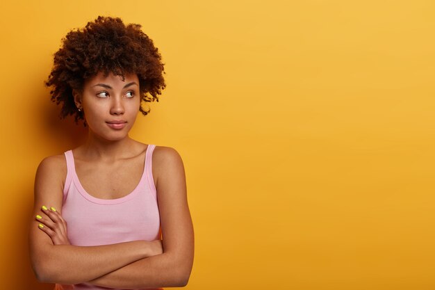 mujer reflexiva tiene una piel sana, cabello rizado natural, mantiene los brazos cruzados y mira pensativamente a un lado, usa ropa casual, aislada sobre una pared amarilla, intenta tomar una decisión