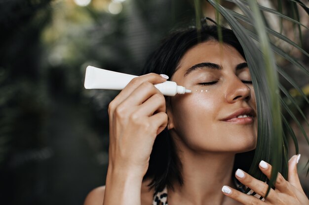 Mujer relajada aplicando protector solar. Mujer alegre con pie de crema facial sobre fondo de naturaleza.