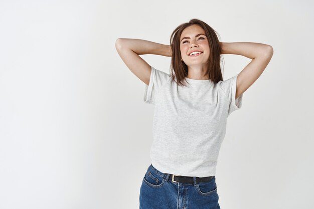 Mujer relajada y feliz descansando y sonriendo, tomados de la mano detrás de la cabeza y mirando hacia arriba con rostro soñador, de pie sin preocupaciones sobre la pared blanca