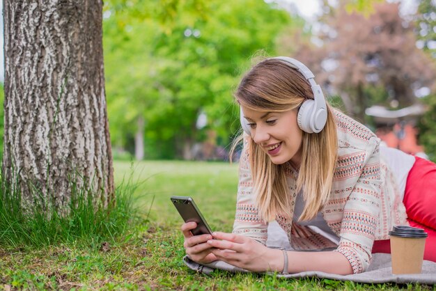 Mujer relajada que escucha la música con los auriculares que mienten en la hierba. Hermosa mujer joven con auriculares disfruta en la música mientras está acostado en la naturaleza, disfrutar de la música