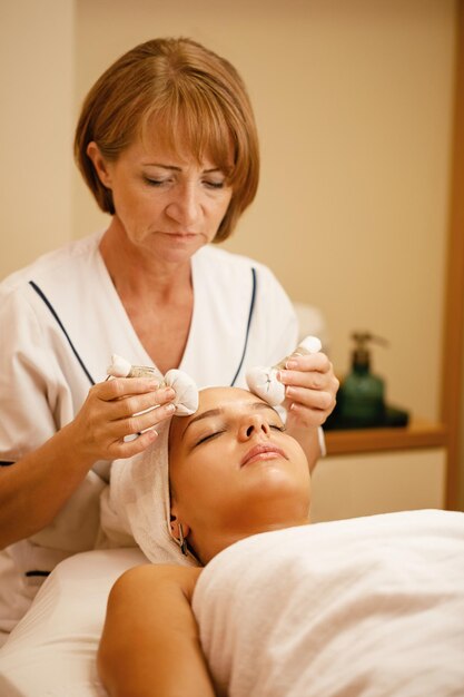 Mujer relajada que recibe masaje facial con bolas de hierbas durante el tratamiento de spa en el salón de belleza