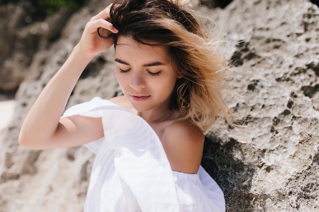 Mujer relajada en vestido blanco vintage de pie en la montaña. Extasiada mujer de cabello oscuro con piel bronceada posando junto a la roca.