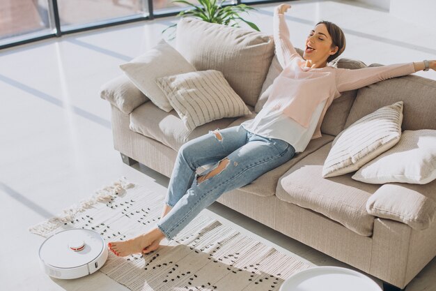 Mujer relajándose en el sofá mientras robot aspirador haciendo quehaceres domésticos