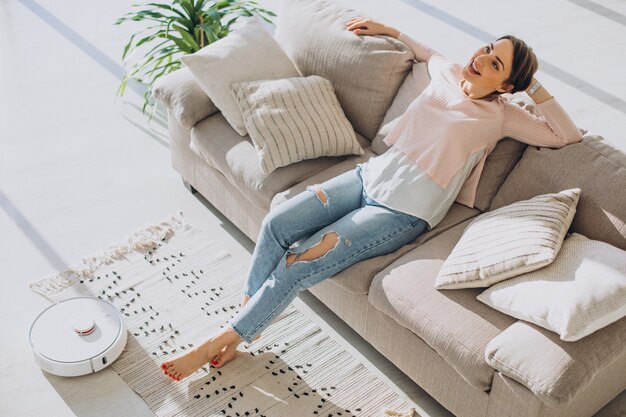 Mujer relajándose en el sofá mientras robot aspirador haciendo quehaceres domésticos