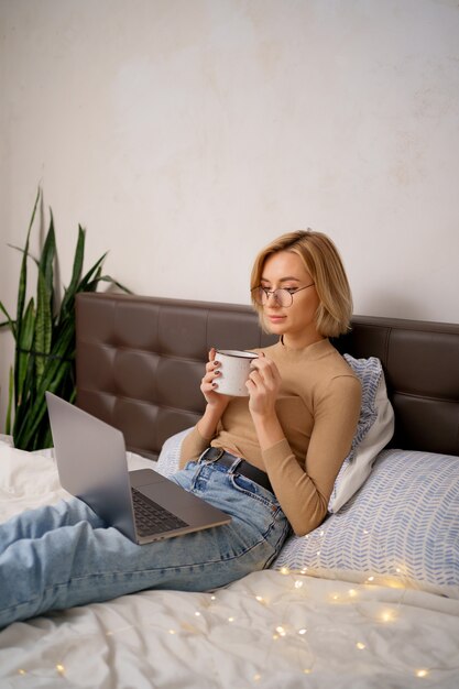 Mujer relajante y bebiendo una taza de té o café caliente usando la computadora portátil en el dormitorio.