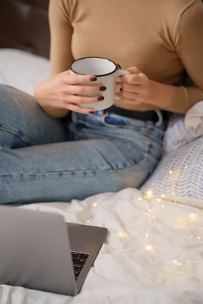 Mujer relajante y bebiendo una taza de té o café caliente usando la computadora portátil en el dormitorio.