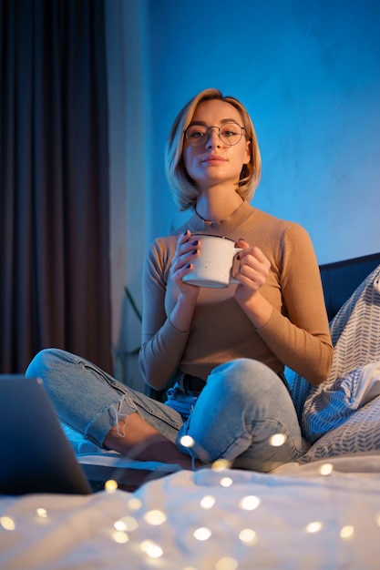 Mujer relajante y bebiendo una taza de té o café caliente usando la computadora portátil en el dormitorio.