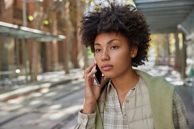 Mujer responde llamada de teléfono inteligente mantiene celular cerca del oído enfocado pensativo hacia adelante vestida con ropa casual plantea afuera durante el día