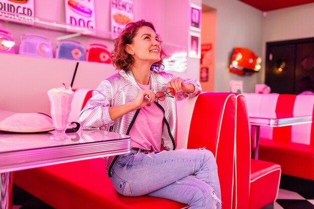 Mujer en retro vintage 50's cafe de color rosa sentado en la mesa bebiendo un cóctel de batido de leche en traje hipster divirtiéndose