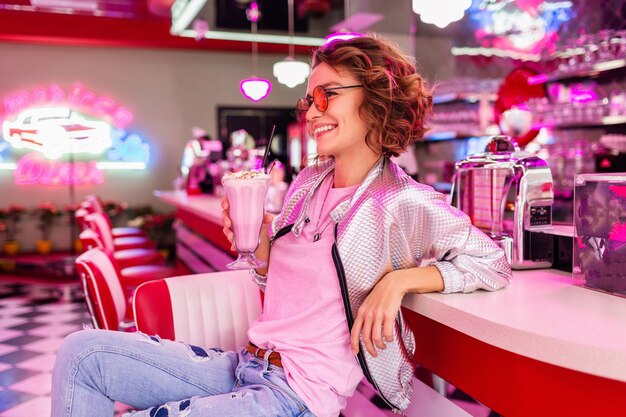 Mujer en retro vintage 50's cafe de color rosa sentado en la mesa bebiendo un cóctel de batido de leche en traje hipster divirtiéndose